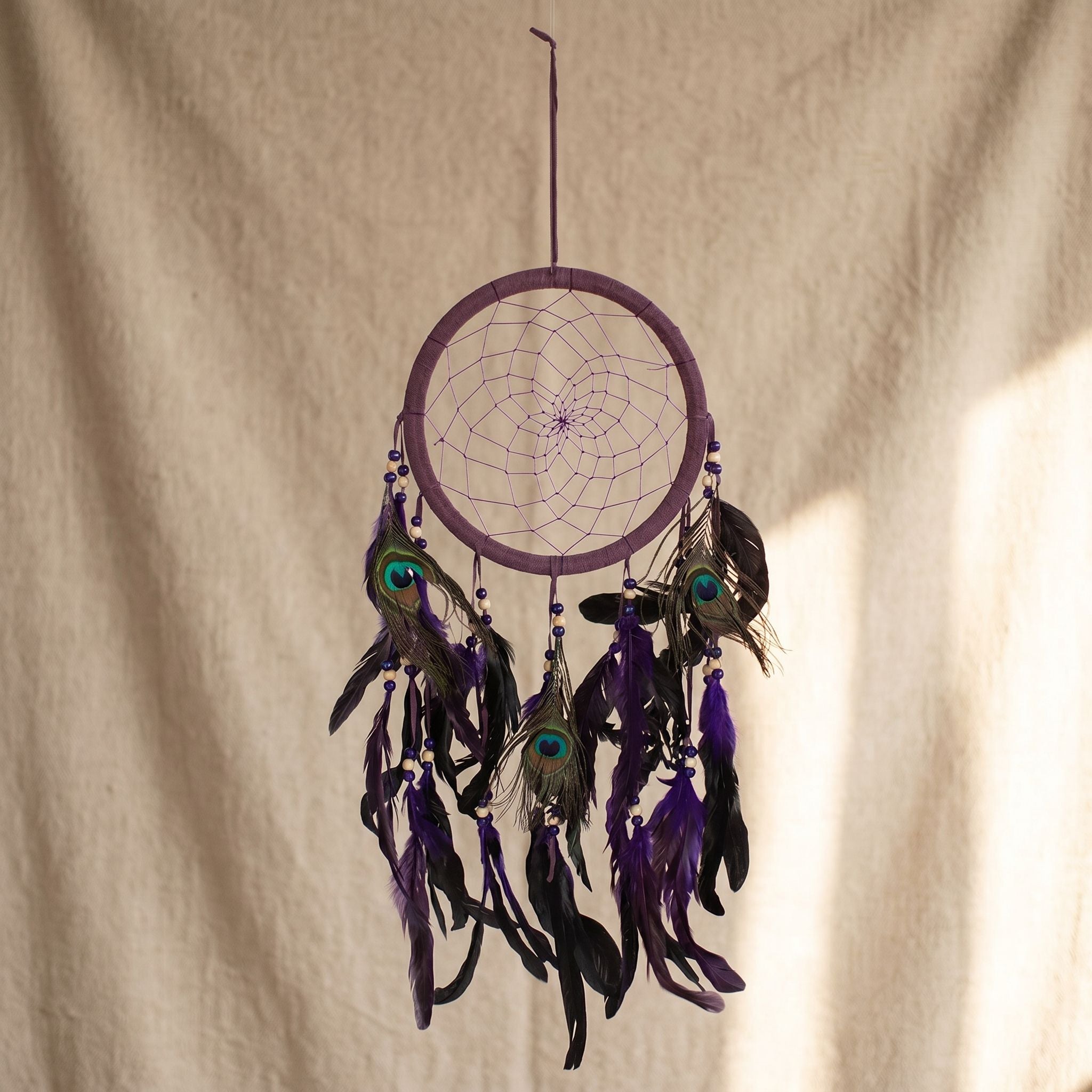 Close-up of purple feather dreamcatcher with peacock feathers and purple stitching against soft, textured tapestry backdrop.